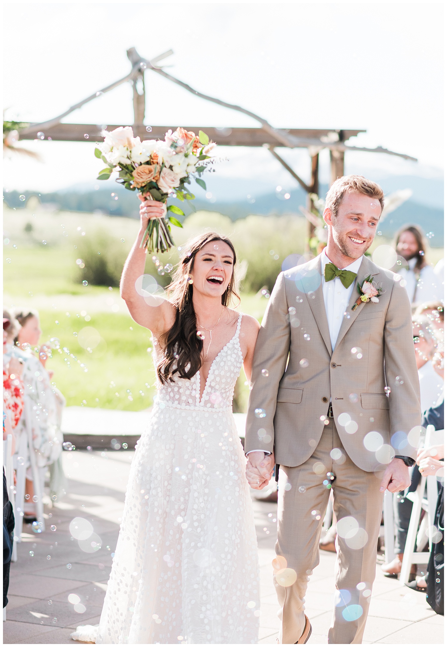 Bride and groom walk back down the aisle celebrating they just got married while bubbles are being blown in their direction and bride raises her bouquet in the air smiling towards the guests all during the best month to get married in Colorado