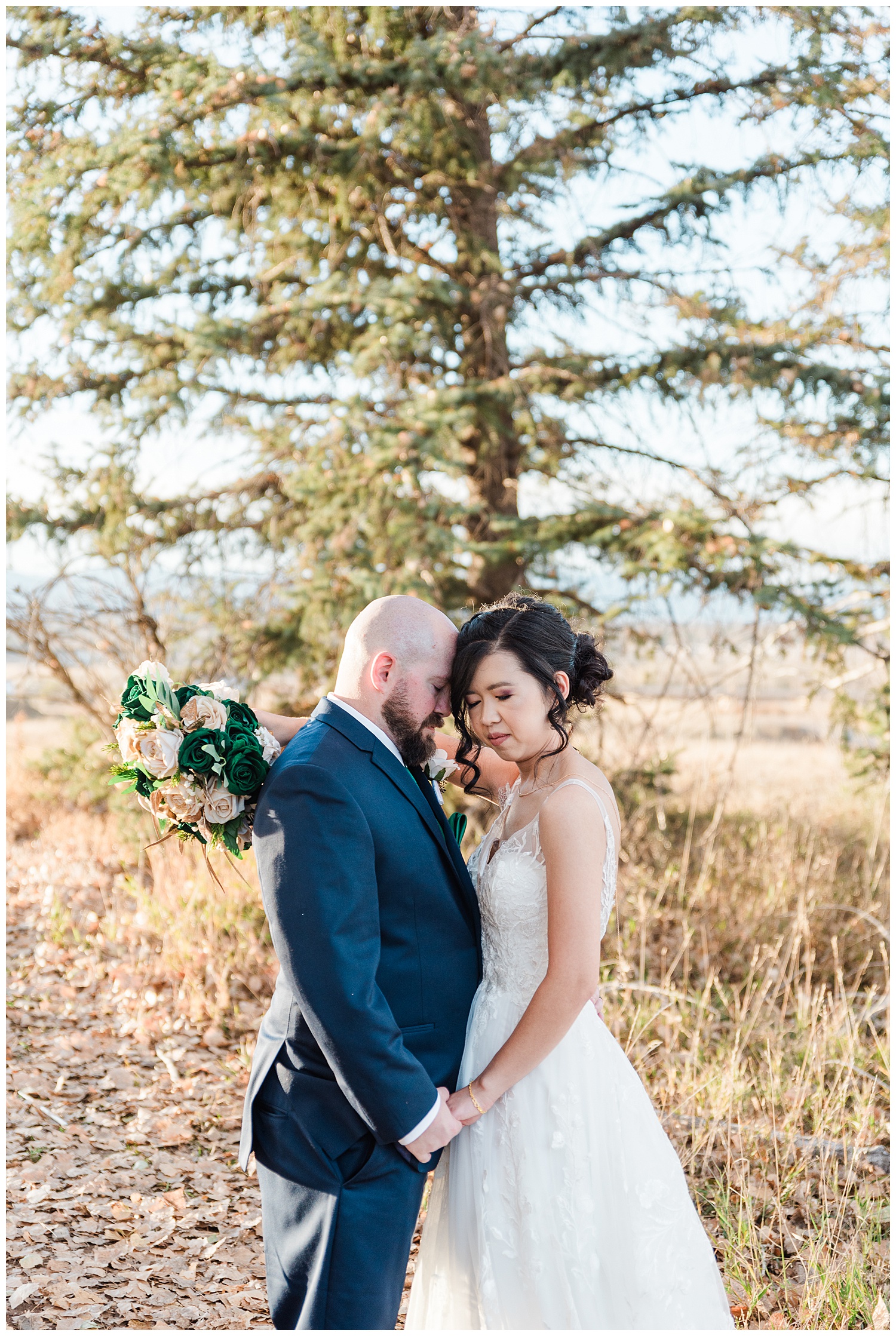 Bride and groom stand in front of a pine tree facing each other while bride looks toward grooms inner elbow and groom places his forehead on her temple 