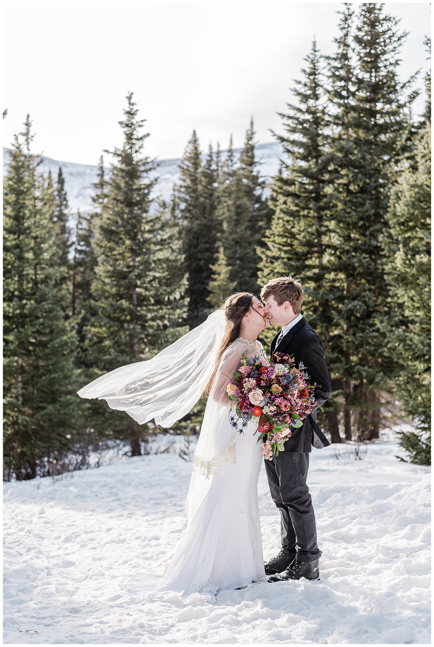 Bride and groom stand on a snowy mountaintop with pine trees behind them and face each other going in for a kiss