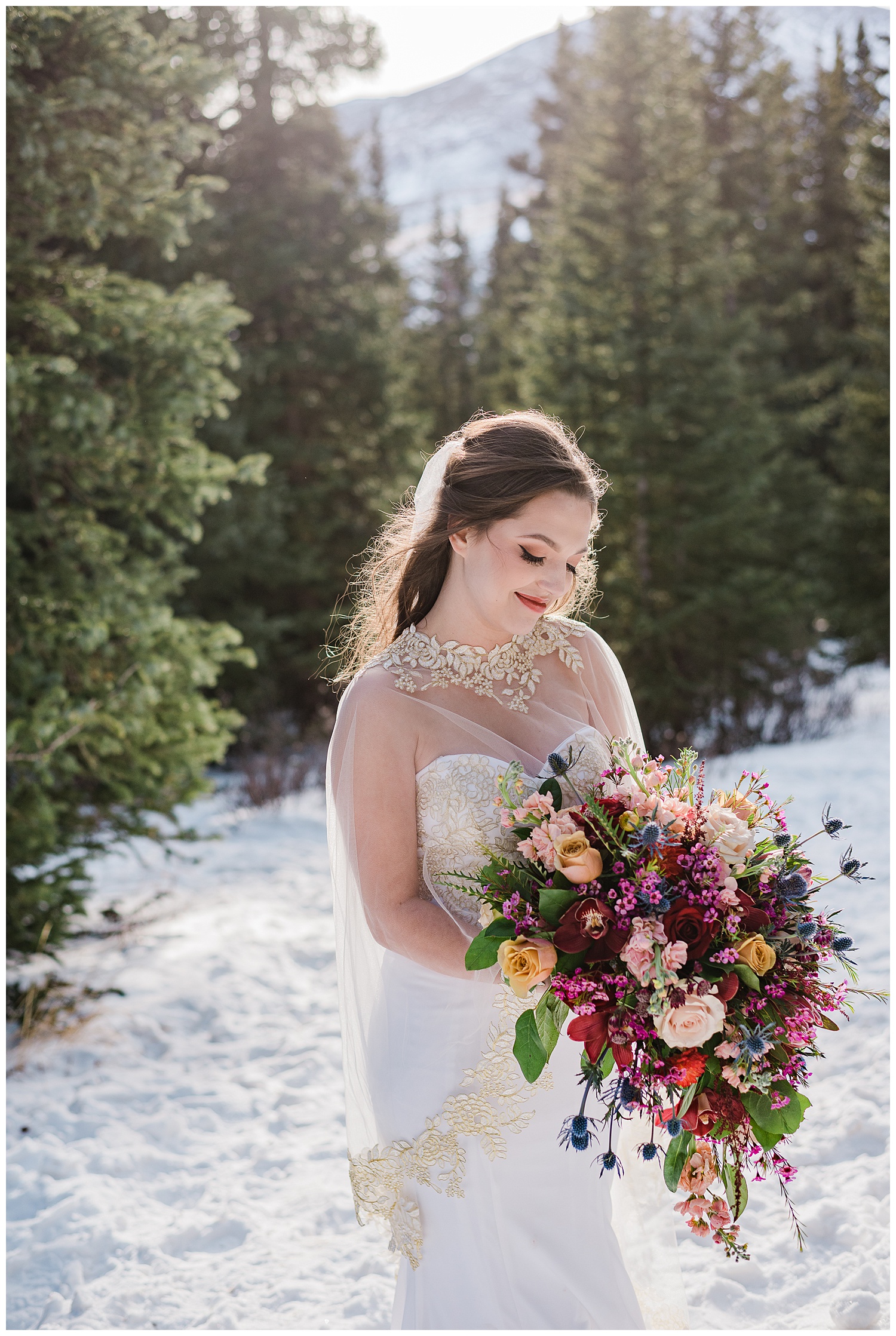 Close up of a bride looking down holding her large and colorful bouquet on a snowy mountain top with pine trees behind her