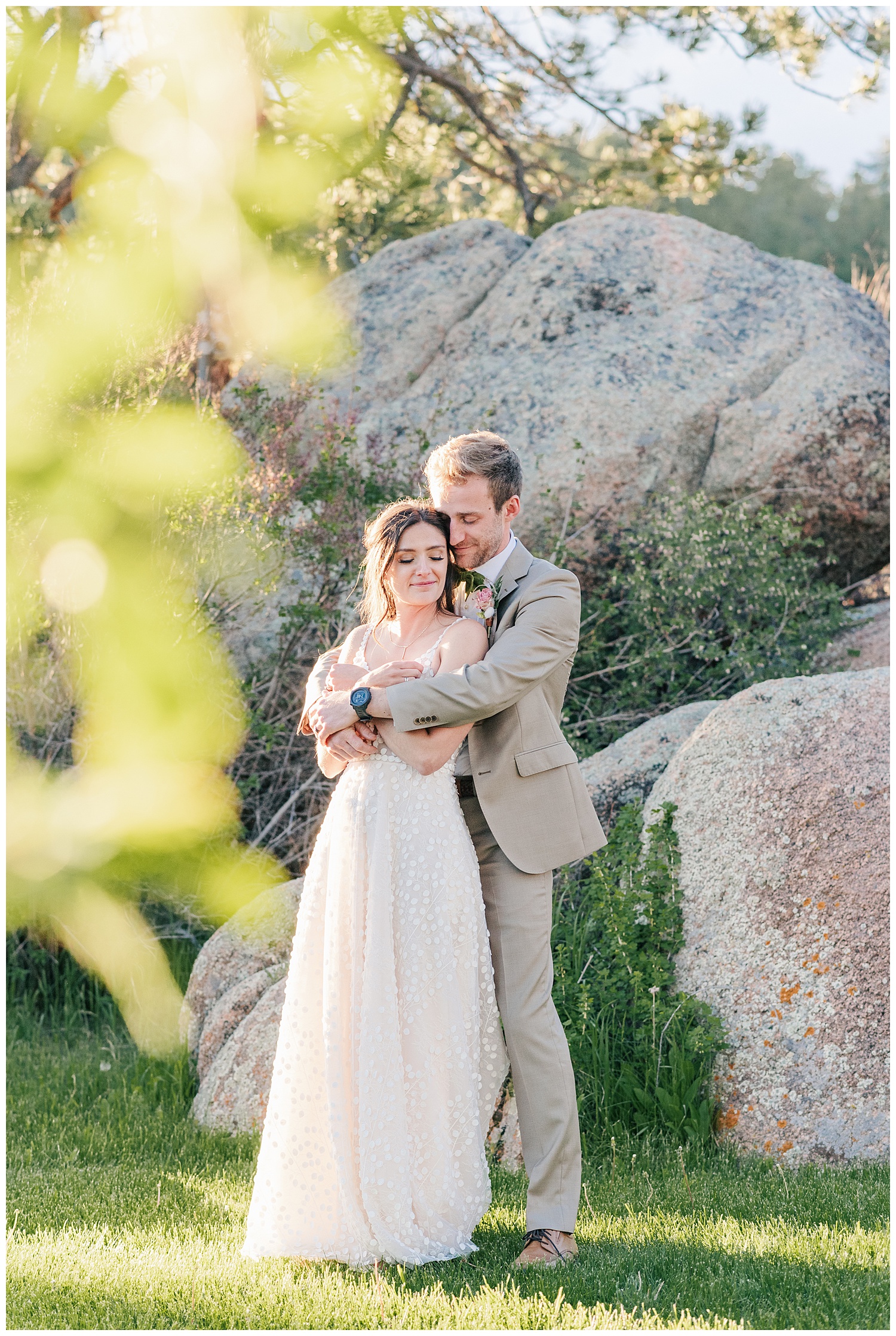 Bride and groom stand together outside in front of large boulders with groom hugging bride from behind as they both smile 