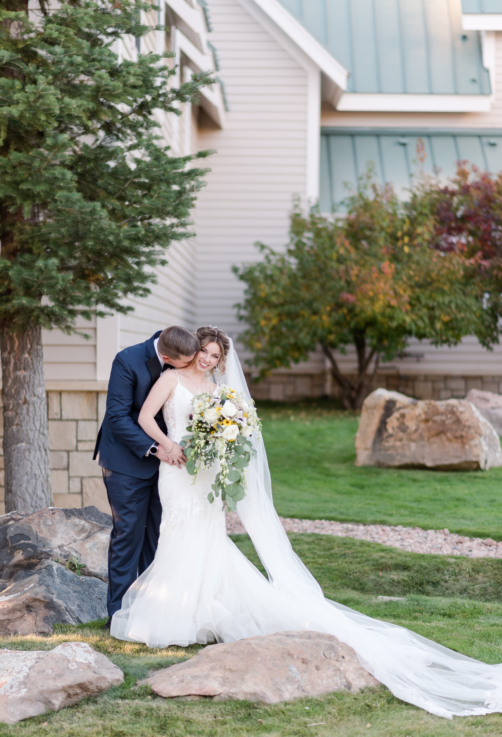 Bride and groom standing together in front of their venue with the groom standing behind the bride kissing her neck and bride smiles at the camera in the best month to get married in Colorado