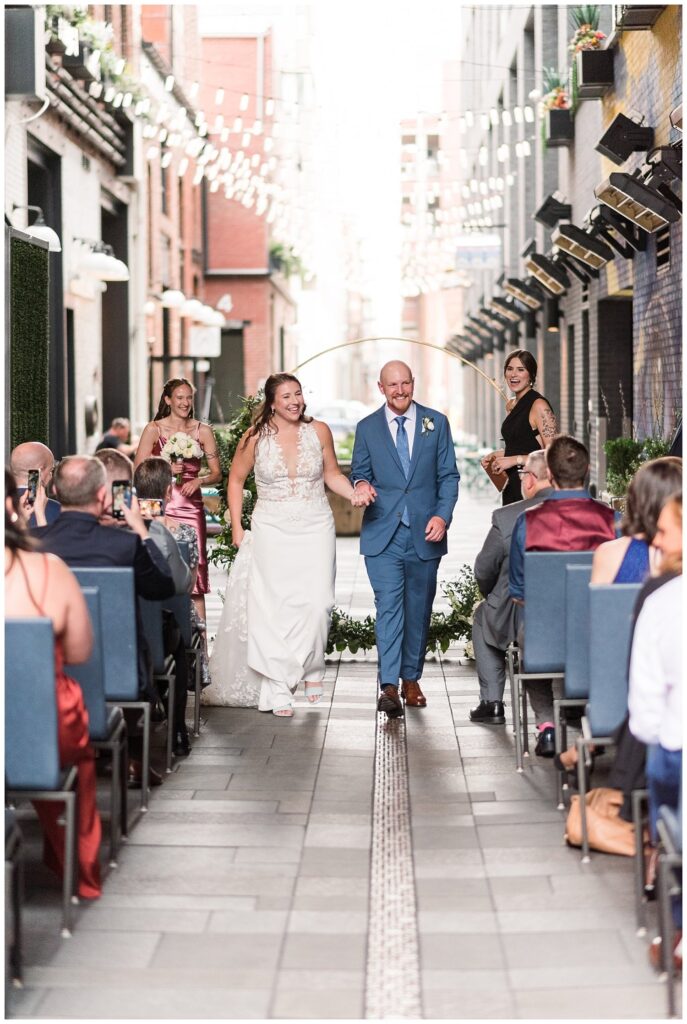 A bride and groom smile and walk down the isle holding hands after their wedding ceremony at The Maven Hotel in Denver with colorado wedding photographers
