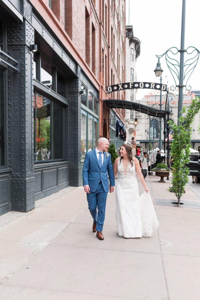 A couple in wedding attire walks down the streets of Denver smiling at each other as a Denver photographer captures their affection for each other