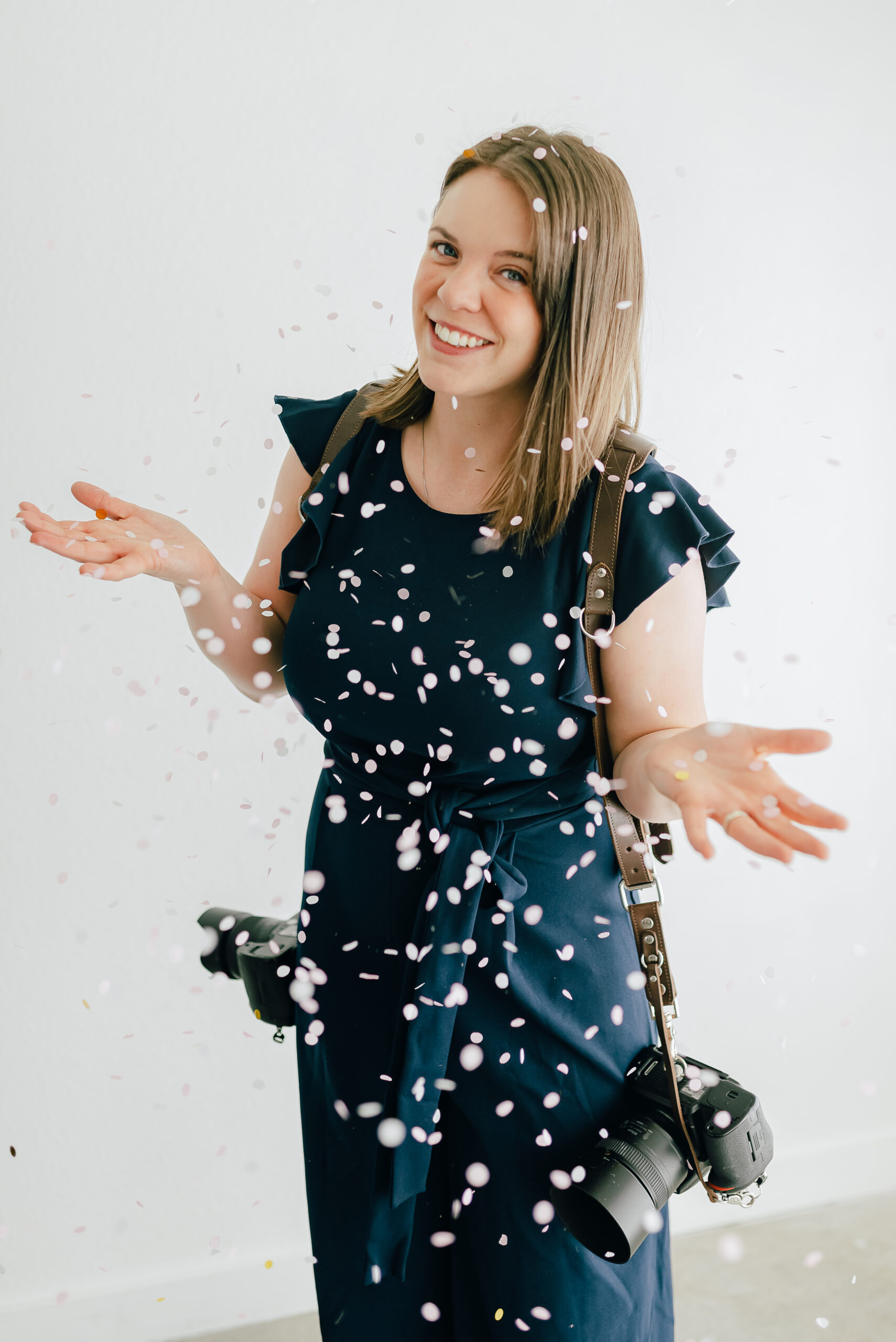 A Denver wedding photographer in a blue jumpsuit with her cameras on each shoulder shrugging with confetti falling down around her.
