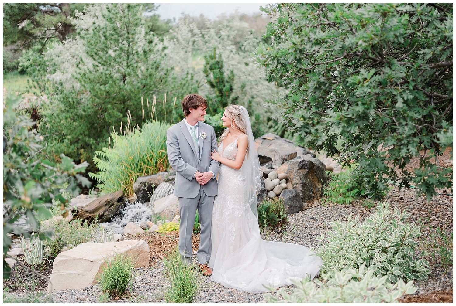 Surrounded by greenery and a waterfall in the background, a bride and groom hold hands while looking at each other and smiling.