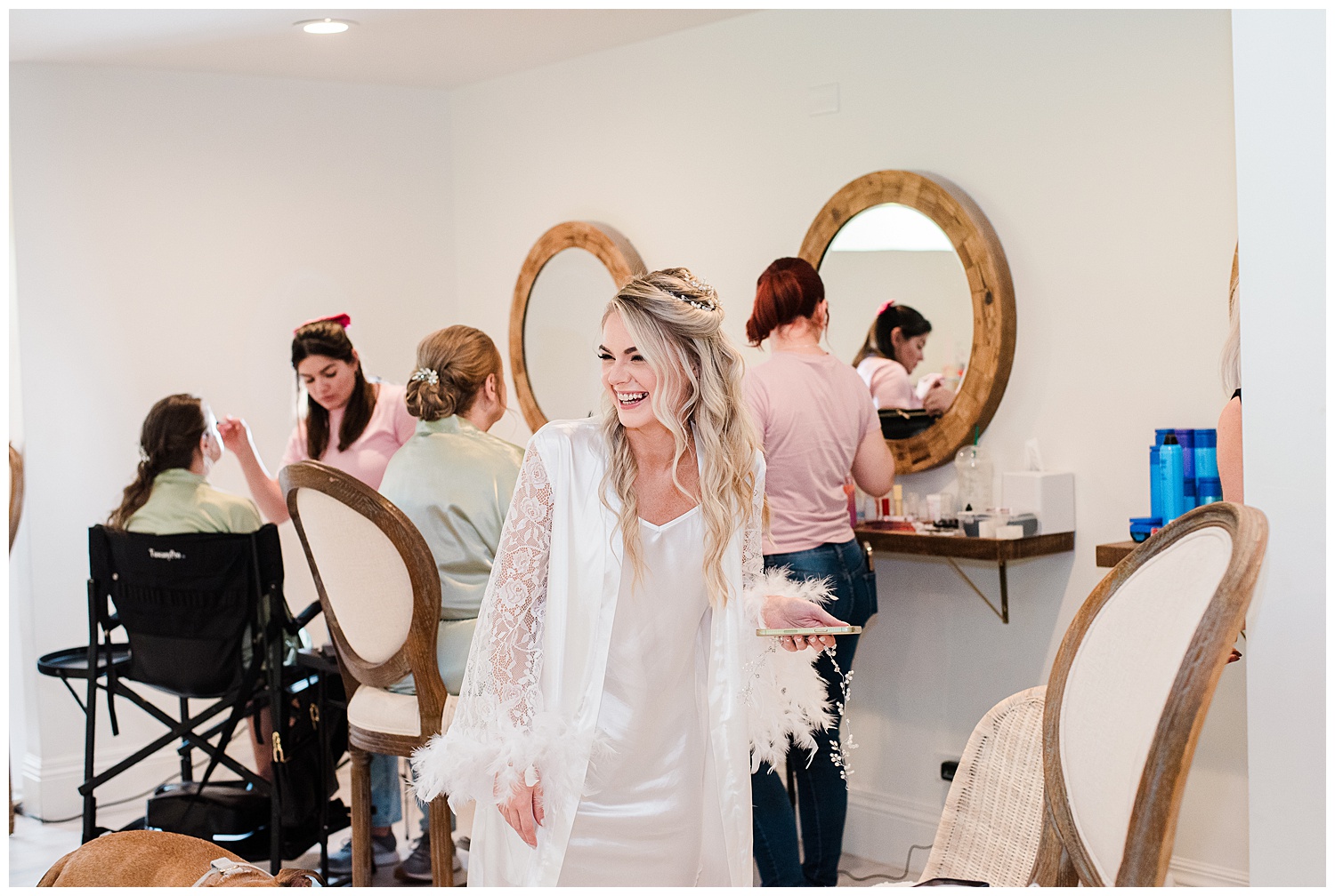 Bride smiles towards her bridesmaids in her getting ready robe while holding her phone in the bridal suite at The Oaks