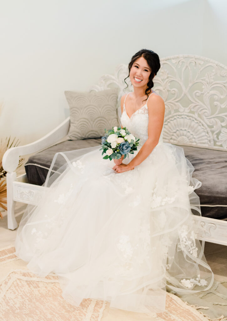 Bride smiles toward the camera sitting on a grey and while couch holding her bouquet in the bridal suite at The Oaks