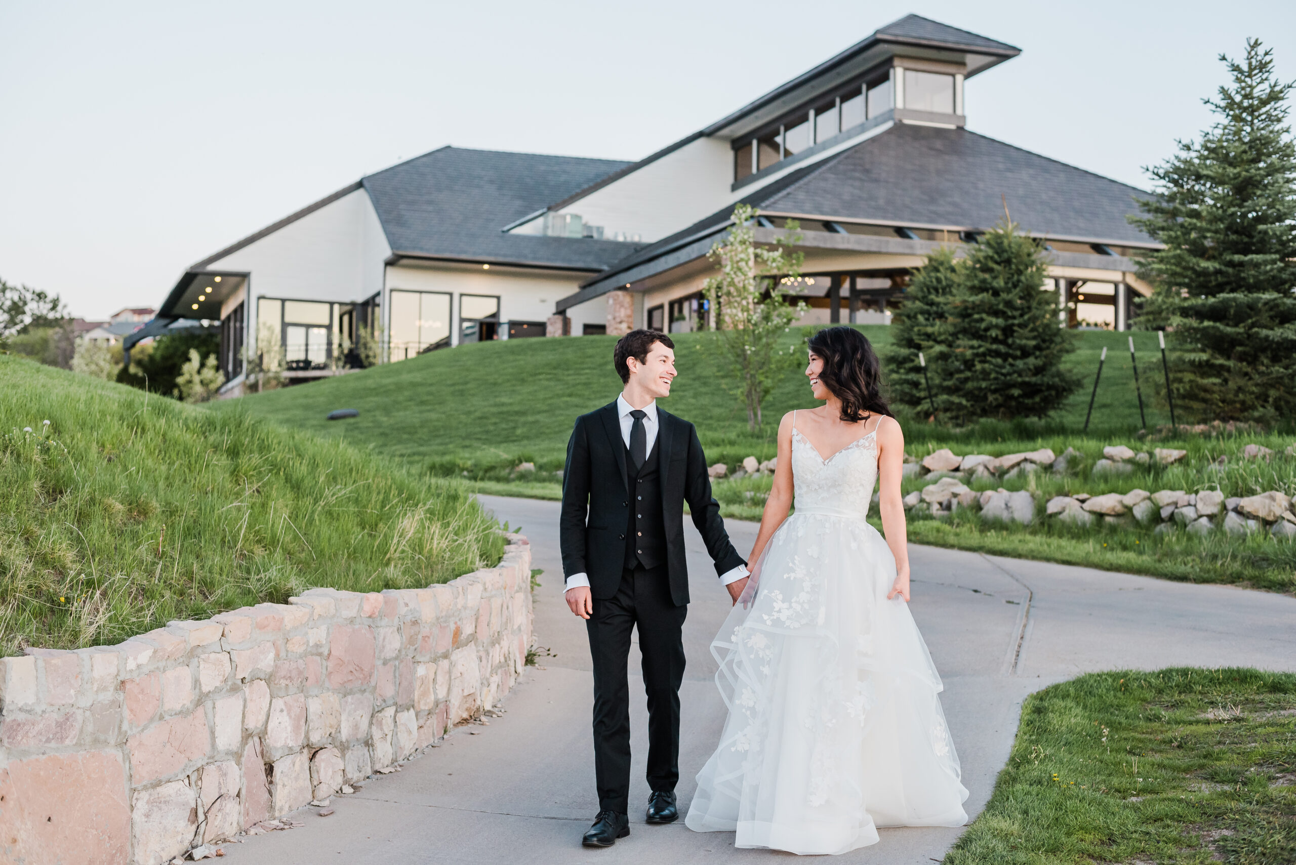 Bride and groom walk hand in hand while looking at each other along a sidewalk path near the wedding venue The Oaks in Castle Rock
