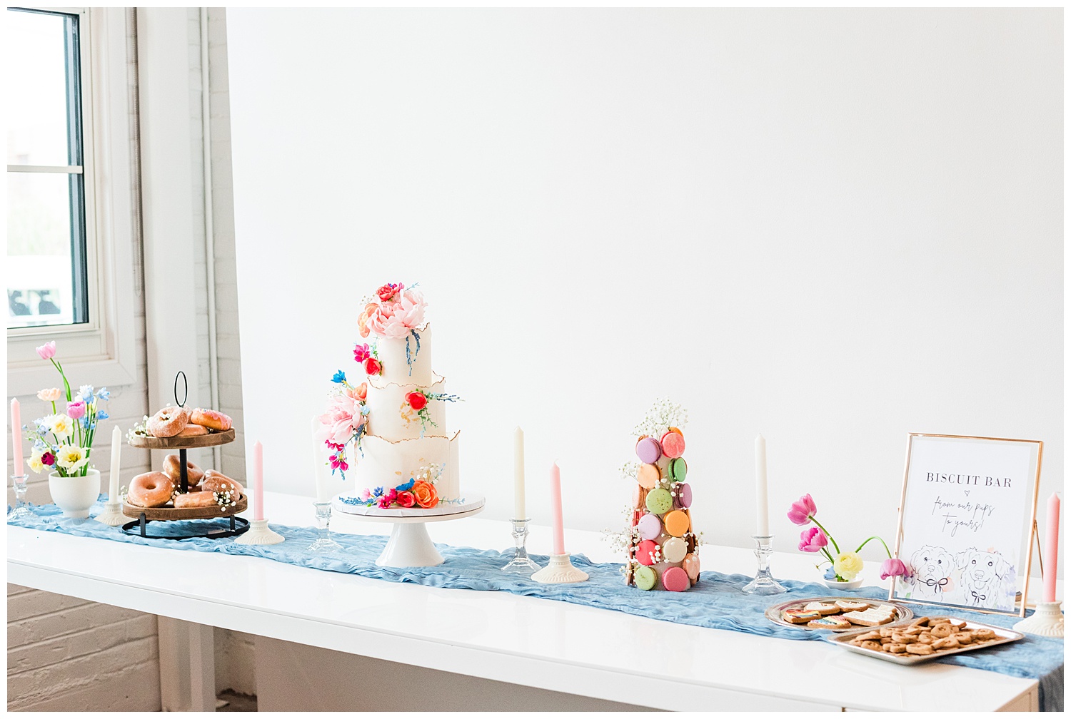 A white dessert table line with a dusty blus cheese cloth and various colorful desserts including a donut tower, a white cake with colorful flowers, a macaroon tower, dog treats, and white candles spread out on the table for decoration
